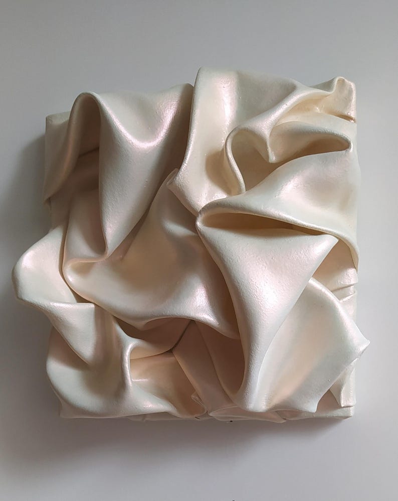 May include: A square, off-white sculpture with a rippled, fabric-like texture. The surface has a subtle pearlescent sheen, catching the light and creating highlights and shadows. The artwork is mounted on a white background.