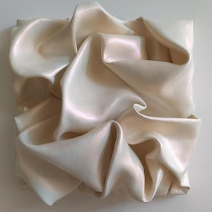 May include: A square, abstract art piece featuring a cream-colored, rippled fabric. The fabric is arranged in a series of folds and curves, creating a dynamic and textured surface. The piece has a subtle sheen, reflecting light and adding depth.