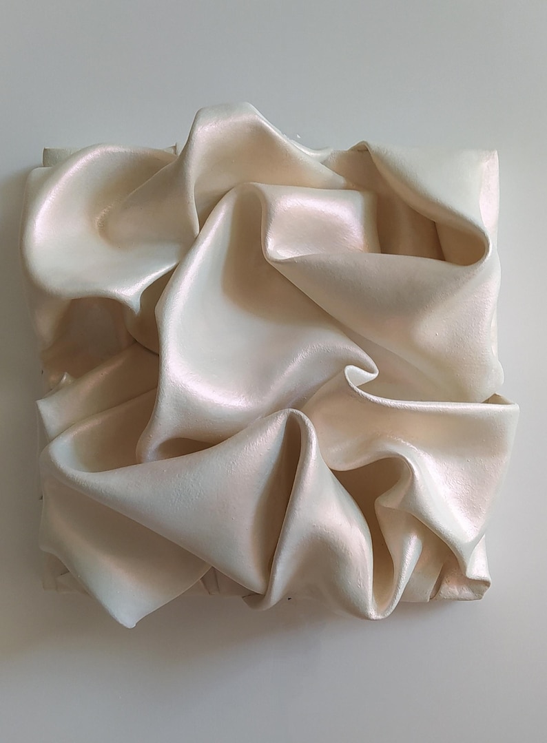 May include: A square, abstract art piece featuring a cream-colored, rippled fabric. The fabric is arranged in a series of folds and curves, creating a dynamic and textured surface. The piece has a subtle sheen, reflecting light and adding depth.