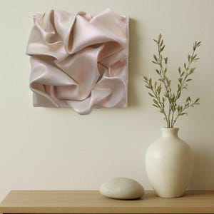 May include: A square, abstract wall art piece with a soft, pinkish-white, rippled fabric design. A white vase with green sprigs and a smooth, gray stone sit on a wooden table. The background is a light beige.