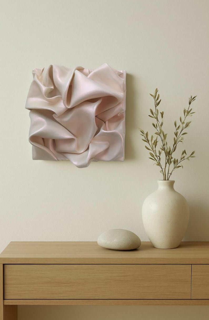 May include: A square, abstract wall art piece with a soft, pinkish-white, rippled fabric design. A white vase with green sprigs and a smooth, gray stone sit on a wooden table. The background is a light beige.