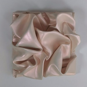 May include: A square, abstract wall art piece featuring a draped, fabric-like design. The piece is primarily a soft, pearlescent pink with highlights and shadows that create a sense of depth and texture. The folds and curves of the material are prominent.