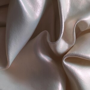 May include: Close-up of a cream-colored, rippled fabric with a pearlescent sheen. The fabric has soft folds and curves, creating a sense of depth and texture. The lighting highlights the subtle color variations.