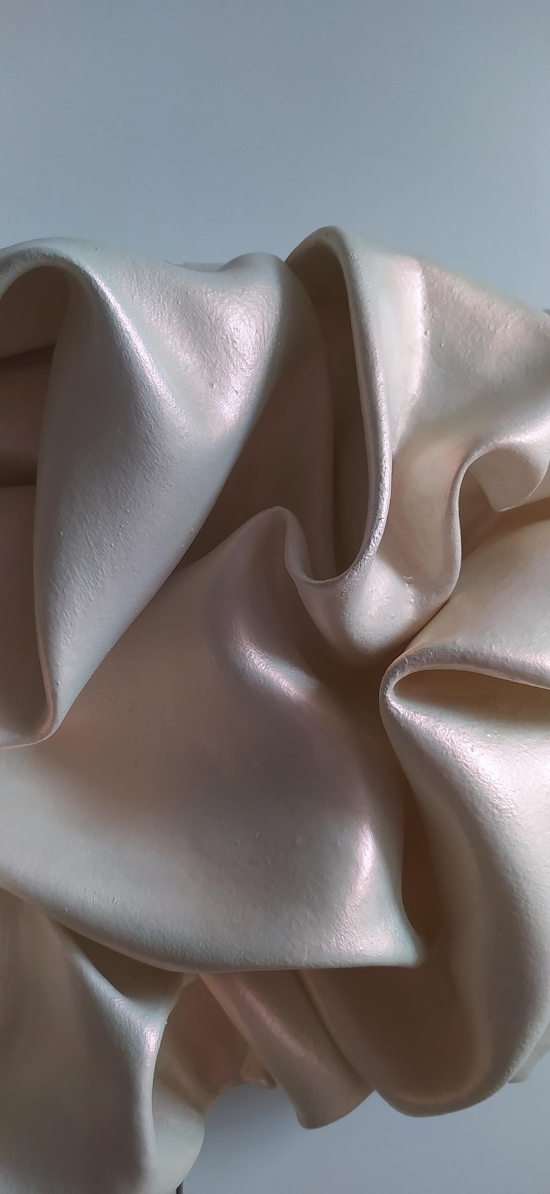 May include: Close-up of a cream-colored, rippled fabric with a pearlescent sheen. The fabric has soft folds and curves, creating a sense of depth and texture. The lighting highlights the subtle color variations.