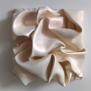 May include: A square, off-white sculpture with a rippled, fabric-like texture. The surface has a pearlescent sheen, with subtle pink and blue highlights. The folds and curves create a dynamic, abstract design, suggesting movement and depth.