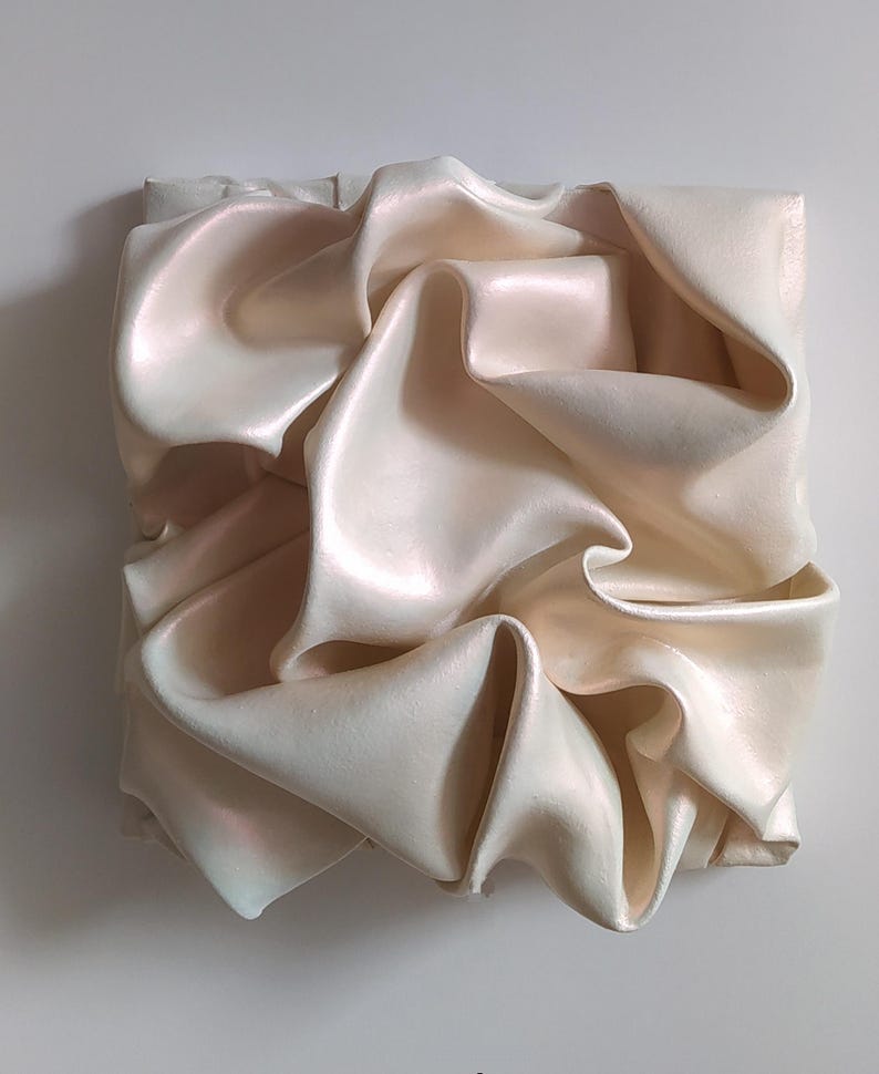 May include: A square, off-white sculpture with a rippled, fabric-like texture. The surface has a pearlescent sheen, with subtle pink and blue highlights. The folds and curves create a dynamic, abstract design, suggesting movement and depth.