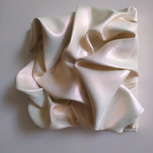 May include: A square, off-white sculpture with a draped fabric design. The fabric appears to be folded and layered, creating a textured surface. The sculpture has a pearlescent sheen, reflecting light.