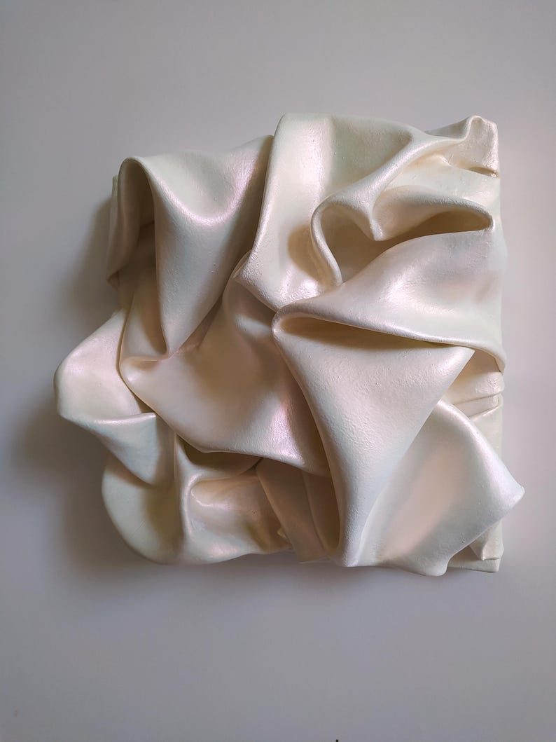 May include: A square, off-white sculpture with a draped fabric design. The fabric appears to be folded and layered, creating a textured surface. The sculpture has a pearlescent sheen, reflecting light.