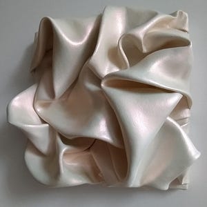 May include: A square, off-white sculpture with a pearlescent finish. The artwork features a draped, fabric-like form with multiple folds and shadows, creating a sense of movement and texture. The piece is mounted on a light gray wall.