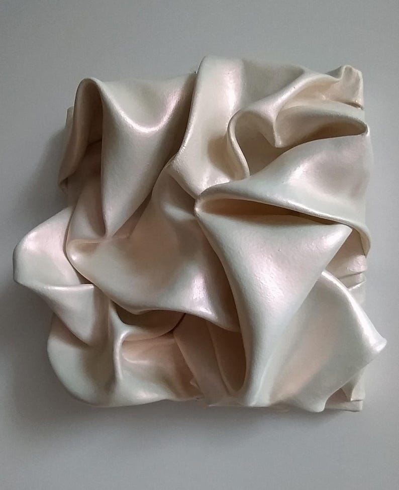 May include: A square, off-white sculpture with a pearlescent finish. The artwork features a draped, fabric-like form with multiple folds and shadows, creating a sense of movement and texture. The piece is mounted on a light gray wall.