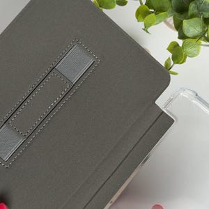 May include: A gray tablet case with a built-in stand and a clear protective cover. The case has a fabric-like texture and a gray elastic strap. The clear cover is made of a transparent material. Green foliage is visible in the background.