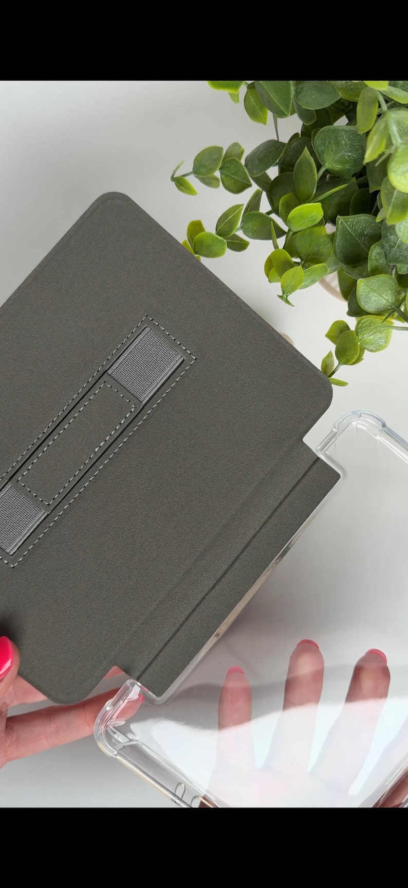 May include: A gray tablet case with a built-in stand and a clear protective cover. The case has a fabric-like texture and a gray elastic strap. The clear cover is made of a transparent material. Green foliage is visible in the background.