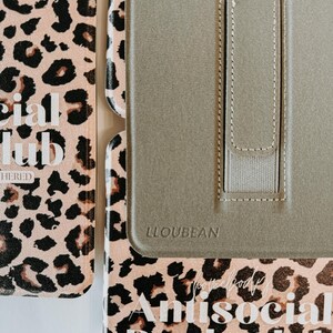 May include: A close-up of a book cover and a tablet case. The book cover has a pink leopard print with the words "Antisocial Book Club" and "Too Booked to be Bothered". The tablet case is olive green with the brand name "LLOUBEAN".