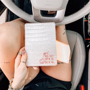 Könnte beinhalten: Weißes, strukturiertes Buchcover mit den roten Worten "girl, READ YOUR BOOK". Das Cover hat ein Krokodilleder-Muster. Das Bild ist aus einem hohen Winkel aufgenommen, wobei das Buch vor einer Person gehalten wird.
