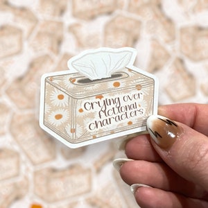 Puede incluir: Una caja de pañuelos blanca con un patrón floral y el texto "Crying over fictional characters" impreso en la parte delantera. La caja está siendo sostenida por una mano con esmalte de uñas marrón y blanco.