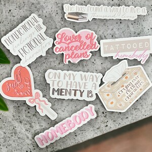 May include: A set of nine stickers with various phrases and designs. The stickers are printed on white paper with pink and brown lettering. The phrases include "Nonfictional feelings for fictional characters", "Lover of cancelled plans", "Suck for smth", "On my way to have a menty b", "Tattooed homebody", "Crying over fictional characters", and "Homebody".