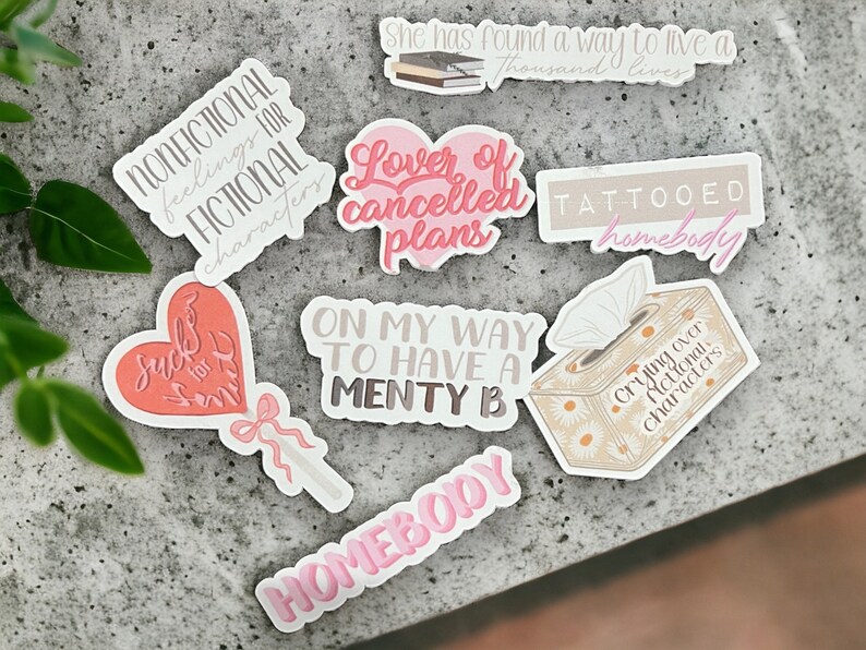 May include: A set of nine stickers with various phrases and designs. The stickers are printed on white paper with pink and brown lettering. The phrases include "Nonfictional feelings for fictional characters", "Lover of cancelled plans", "Suck for smth", "On my way to have a menty b", "Tattooed homebody", "Crying over fictional characters", and "Homebody".