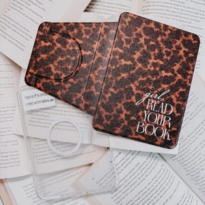Op de afbeelding: Een bruin en zwart telefoonhoesje met luipaardprint met de tekst "girl read your book" in het wit. De hoes is open en onthult een doorzichtig telefoonhoesje. De items liggen op open boeken.