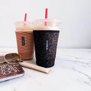May include: Two iced coffee cups with sleeves, one featuring a brown and white horizontal stripe pattern and the other a black and brown leopard print. Both sleeves display the vertical text H.LOUBEAN. Each cup includes a red plastic straw.