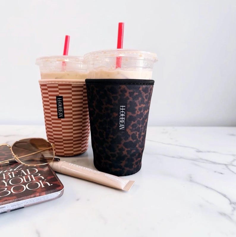 May include: Two iced coffee cups with sleeves, one featuring a brown and white horizontal stripe pattern and the other a black and brown leopard print. Both sleeves display the vertical text H.LOUBEAN. Each cup includes a red plastic straw.