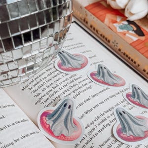 May include: A collection of ghost-shaped stickers, each featuring a gray ghost floating on a pink inner tube. The stickers are placed on the pages of an open book. A disco ball is in the background.