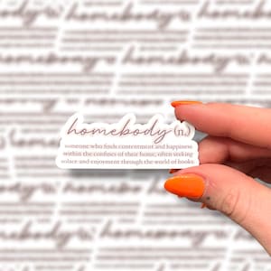 May include: A rectangular sticker with the word "homebody" in a cursive font, with a definition below. The sticker is held by a hand with orange nail polish. The background has a repeating pattern of the word "homebody".