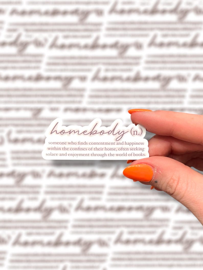A hand with bright orange nails holds a sticker that reads "homebody (n.): someone who finds contentment and happiness within the confines of their home, often seeking solace and enjoyment through the world of books." The background features a blurred, repeated pattern of the same design.