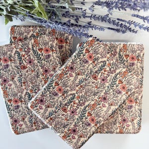May include: Set of four floral print coasters. The coasters feature a cream background with a pattern of pink, orange, and purple flowers and green leaves. The coasters are square with a white border. A bundle of lavender is in the background.
