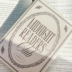 May include: A light-colored rectangular card with the text "MIDNIGHT READERS CLUB" in a decorative font, surrounded by floral embellishments. The card also includes the text "MEMBERS ONLY" and "AFTER DARK". The card is resting on top of an open book.