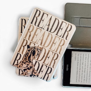 Puede incluir: Una funda protectora para lector electrónico con un patrón repetitivo en color marrón de la palabra READER y un gráfico de un leopardo. La funda está sobre un lector electrónico negro con una pantalla que muestra texto y la palabra kindle.