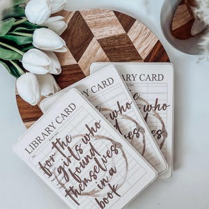 May include: Three library cards with the text "for the girls who found themselves in a book" and coffee ring stains. The cards are on a wooden board with a diamond pattern, surrounded by white tulips and a glass.