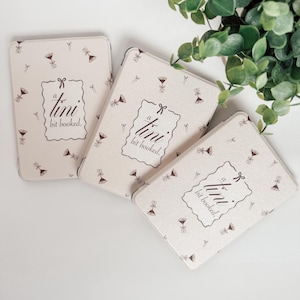 May include: Three rectangular, off-white tins with a repeating pattern of small martini glasses and bows. Each tin features the text "a tini bit booked" in a decorative frame. The tins are arranged on a white surface with green foliage in the background.