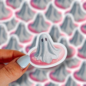 May include: A sticker featuring a gray ghost with black eyes, floating on a pink inner tube. The sticker is held by a hand with a light blue painted fingernail. The background shows a pattern of similar ghost stickers.