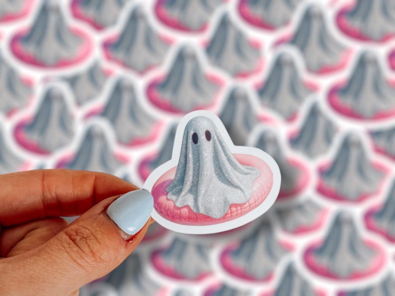 May include: A sticker featuring a gray ghost with black eyes, floating on a pink inner tube. The sticker is held by a hand with a light blue painted fingernail. The background shows a pattern of similar ghost stickers.