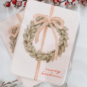 May include: Two rectangular cards with a watercolor design of a green wreath and a pink bow. The cards have the text "merry bookmas" in red. The background is white.