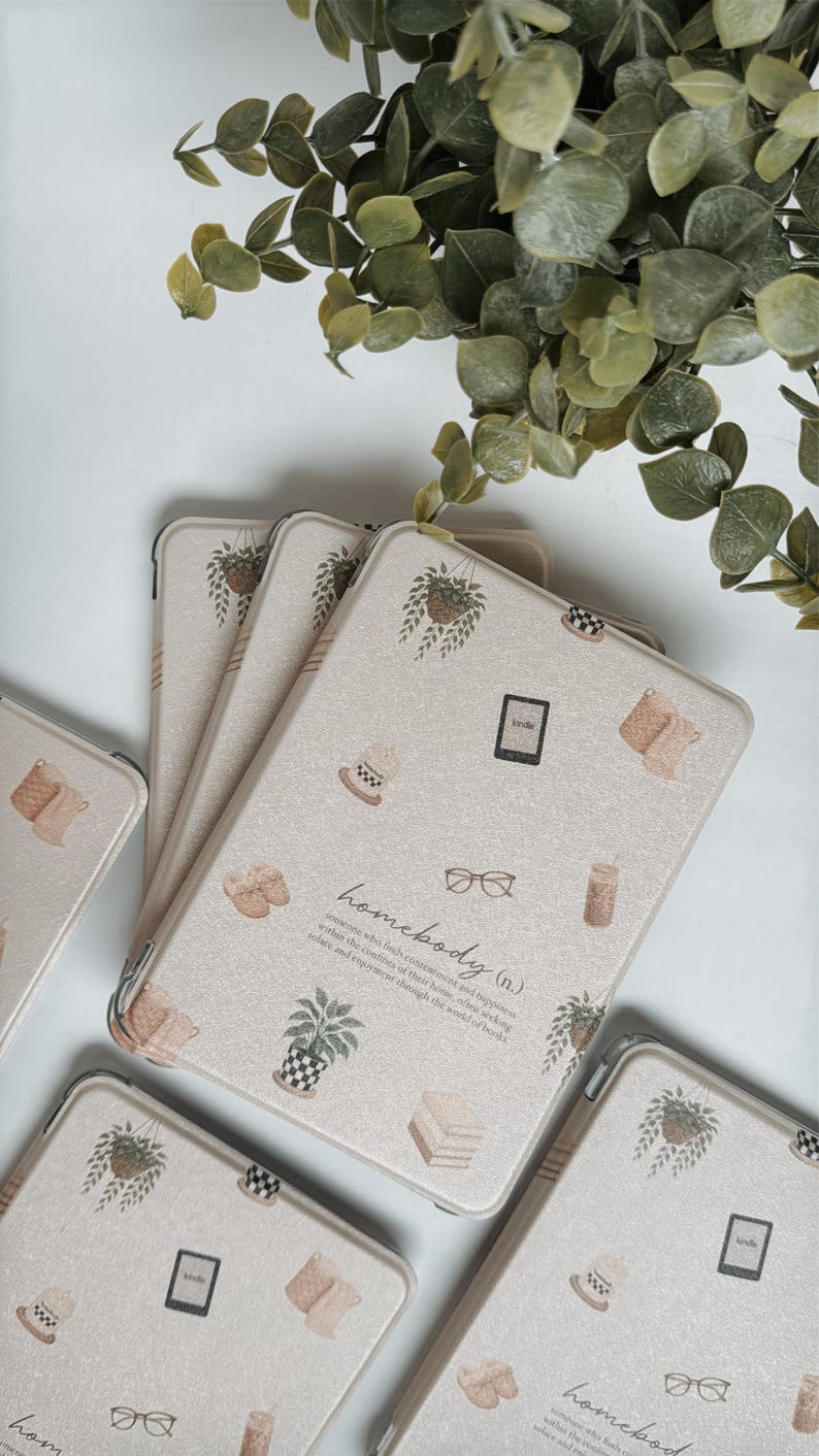 May include: Beige Kindle covers with a repeating pattern of potted plants, books, and other home-themed illustrations. The word "homebody" is printed on the cover. Green leafy plant stems are in the background.