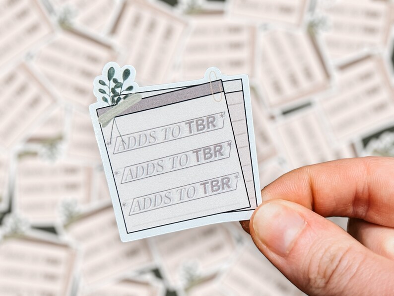 A hand holds a sticker designed like a notepad with "Adds to TBR" written three times in a typewriter-style font. The sticker has decorative elements, including a small green plant, a paperclip, and soft neutral tones, with a blurred background of similar stickers scattered behind it.