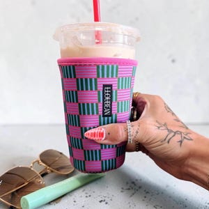 May include: A neoprene iced coffee cup sleeve featuring a checkered pattern of pink and teal stripes. The sleeve has a black rectangular label with the white text LLOUBEAN. It is placed around a clear plastic cup with a red straw and light brown liquid.