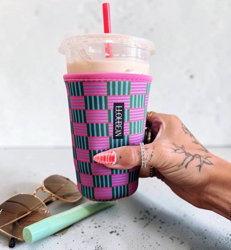 May include: A neoprene iced coffee cup sleeve featuring a checkered pattern of pink and teal stripes. The sleeve has a black rectangular label with the white text LLOUBEAN. It is placed around a clear plastic cup with a red straw and light brown liquid.