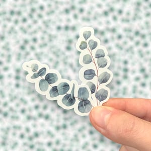 May include: Two watercolor eucalyptus sprigs with green and blue leaves on a white background. The sprigs are arranged in a V-shape.