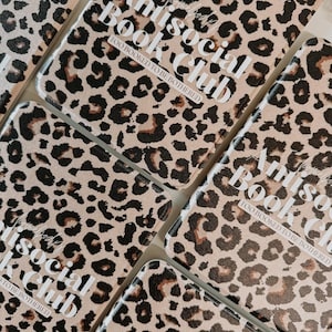 May include: Several rectangular book covers with a tan leopard print pattern. The covers have the text "Antisocial Book Club" and "Too Booked to be Bothered" in white lettering. The covers are arranged in a close-up shot.
