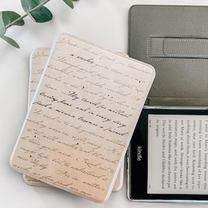 Puede incluir: Una funda para lector electrónico de color beige con texto escrito a mano en cursiva negra sobre un fondo marrón claro. La funda está junto a un lector electrónico negro con texto visible en la pantalla y una funda protectora gris.