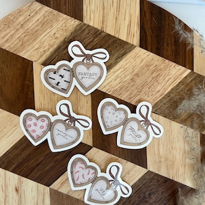 Op de afbeelding: Hartvormige stickers met een medaillonontwerp. Elke sticker heeft een ander patroon aan de binnenkant, waaronder vleermuizen, lippen en aardbeien. De tekst op de stickers luidt "Fantasy girlie", "Romance girlie", "Thriller girlie" en "Smut girlie."