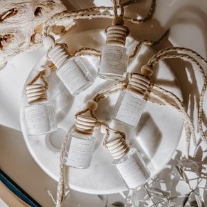 May include: Six clear glass diffuser bottles with wooden tops and hanging cords are arranged on a white marble tray. Each bottle has a label with text. The bottles are surrounded by decorative elements, including a piece of driftwood and a book.