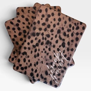 May include: A stack of three rectangular book covers featuring a tan background with a black leopard print pattern. The top cover displays the text "book lover" in a white, cursive, handwritten-style font angled across the bottom right corner.