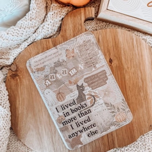 May include: A rectangular decorative coaster with a collage design featuring text, pumpkins, a black cat, and a skeleton. The coaster reads "I lived in books more than I lived anywhere else."