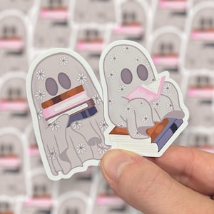May include: Two white ghost stickers with floral patterns. One ghost is holding a stack of books and the other is reading a pink book. Both ghosts are sitting on top of books.