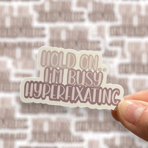 May include: A white sticker with a pink and brown gradient design. The sticker says "Hold on. I'm busy hyperfixating."