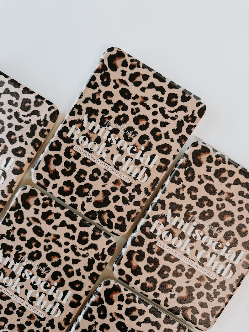 May include: Several rectangular leopard print book covers are arranged on a white surface. The covers feature a tan background with dark brown spots. White text on each cover reads "Antisocial Book Club, Too Booked To Be Bothered."