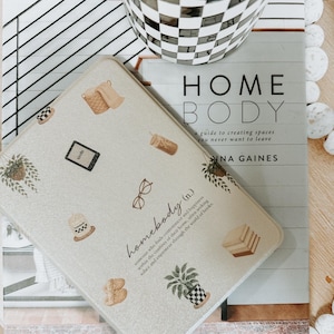 May include: A decorative, light-colored tray with various illustrations, including a potted plant, glasses, and a coffee cup. The tray is on top of a book titled "HOME BODY" and a black and white checkered mug.